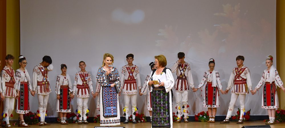 Daniela Toma alături de Ioana Anton, prezentatoarea spectacolului, pe scena Filarmonicii Pitești, cu Ansamblul „Mugurelul” din Costești în fundal